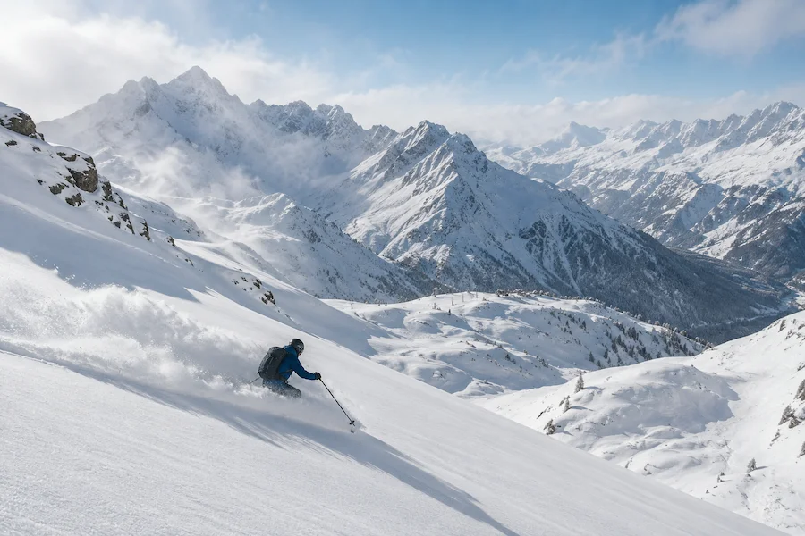 Esquiador descendiendo sobre nieve polvo en una ladera de los Alpes, con un gran paisaje de montañas nevadas al fondo.