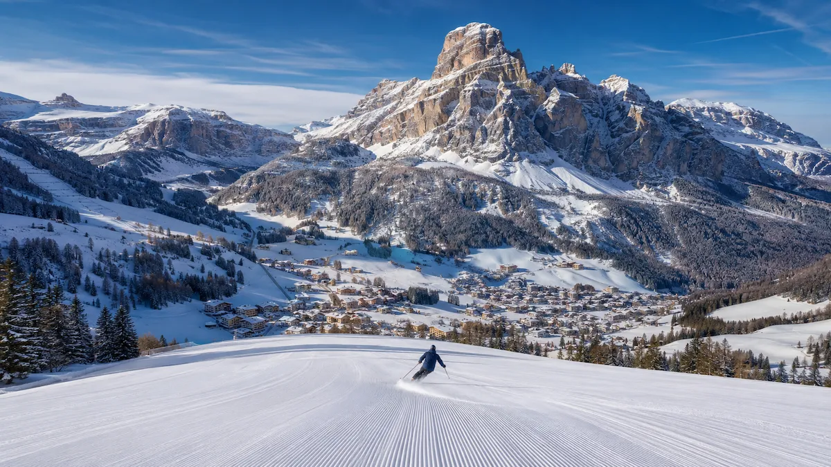 Esquiador descendiendo una pista amplia en Alta Badia, con Corvara y las montañas de los Dolomitas al fondo.