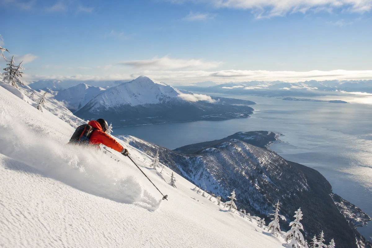 Esquiador descendiendo sobre nieve polvo en Alyeska con vistas al océano y las montañas costeras de Alaska al atardecer.