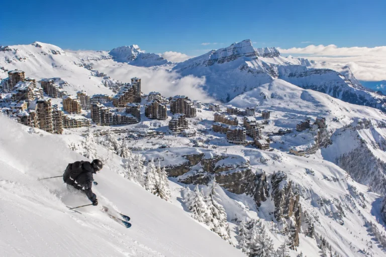 Esquiador descendiendo sobre Avoriaz con la estación nevada en altura, arquitectura integrada y montañas de Portes du Soleil al fondo.