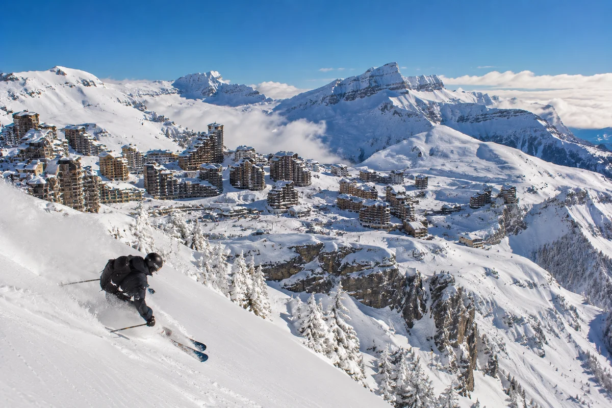 Esquiador descendiendo sobre Avoriaz con la estación nevada en altura, arquitectura integrada y montañas de Portes du Soleil al fondo.