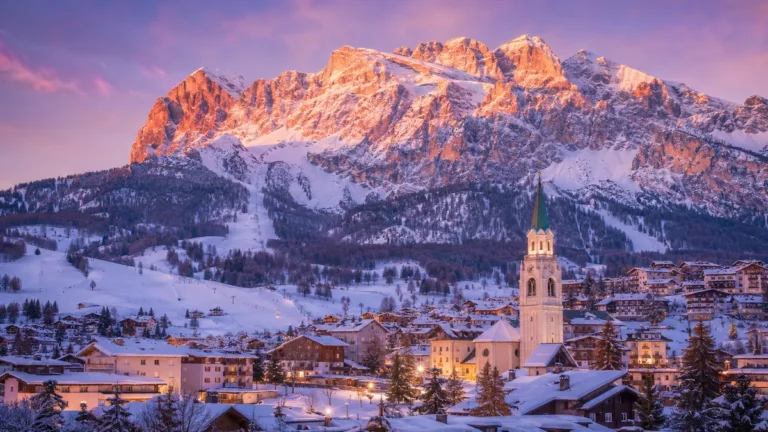 Cortina d’Ampezzo nevada al atardecer, con el campanario del pueblo y las montañas Dolomitas iluminadas por el alpenglow.