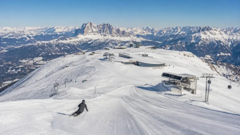 Esquiador descendiendo por una pista amplia en Kronplatz, con remontes modernos en la cima y los Dolomitas al fondo.