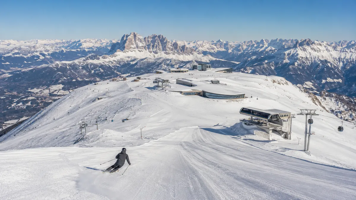Esquiador descendiendo por una pista amplia en Kronplatz, con remontes modernos en la cima y los Dolomitas al fondo.