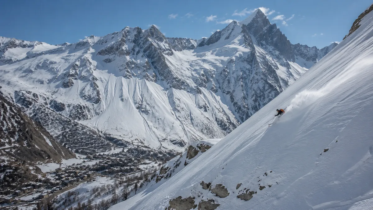 Esquiador descendiendo una ladera muy inclinada en La Grave con el pueblo abajo y grandes cumbres alpinas al fondo