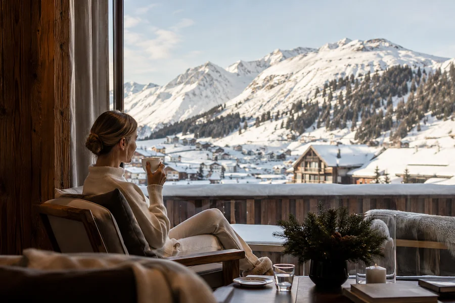 Interior de hotel alpino en Lech Zürs con huésped contemplando el pueblo nevado y las montañas desde una ventana panorámica