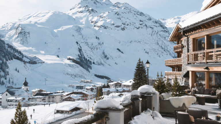 Chalet alpino de lujo en Lech Zürs con terraza nevada, pueblo austríaco y montañas al fondo en un día luminoso