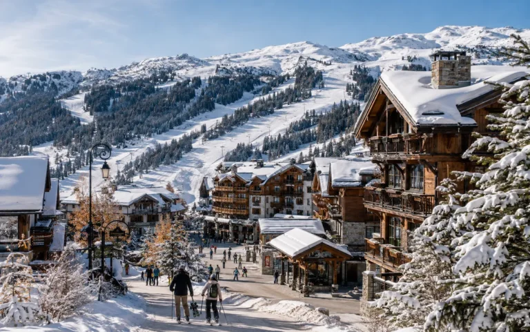 Pueblo alpino de Méribel cubierto de nieve, con chalets de madera, esquiadores y montañas de Les Trois Vallées al fondo.