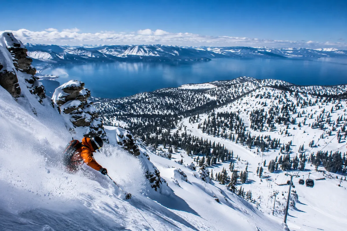 Esquiador descendiendo entre roca y nieve en Palisades Tahoe con vistas panorámicas al lago Tahoe bajo cielo despejado.