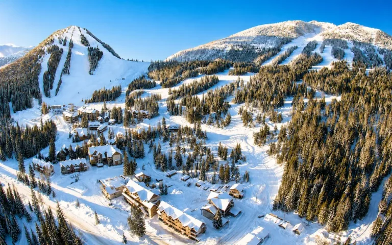 Vista aérea soleada de Red Mountain en British Columbia, con pistas entre bosque, nieve abundante y alojamientos a pie de pista.