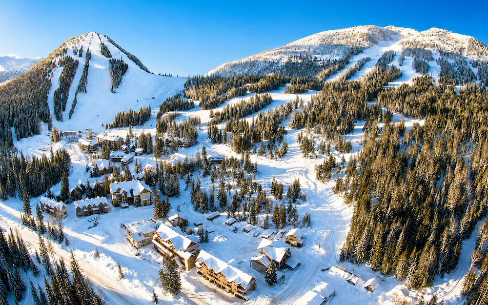Vista aérea soleada de Red Mountain en British Columbia, con pistas entre bosque, nieve abundante y alojamientos a pie de pista.