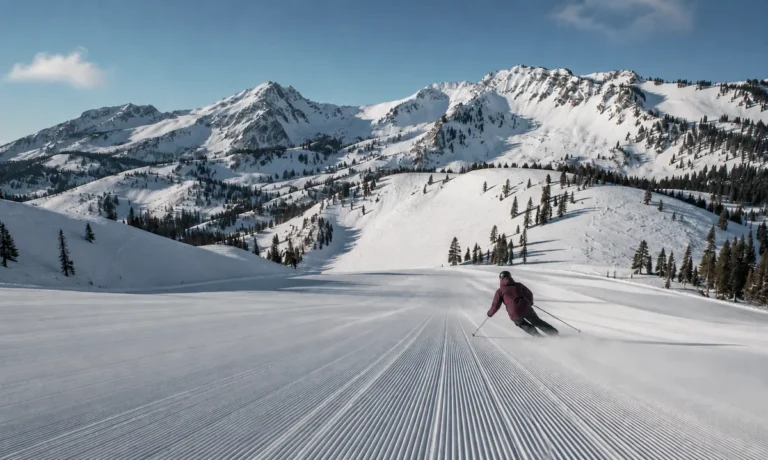 Esquiador descendiendo por una pista amplia y recién pisada en Snowbasin, Utah, con montañas nevadas y terreno olímpico al fondo