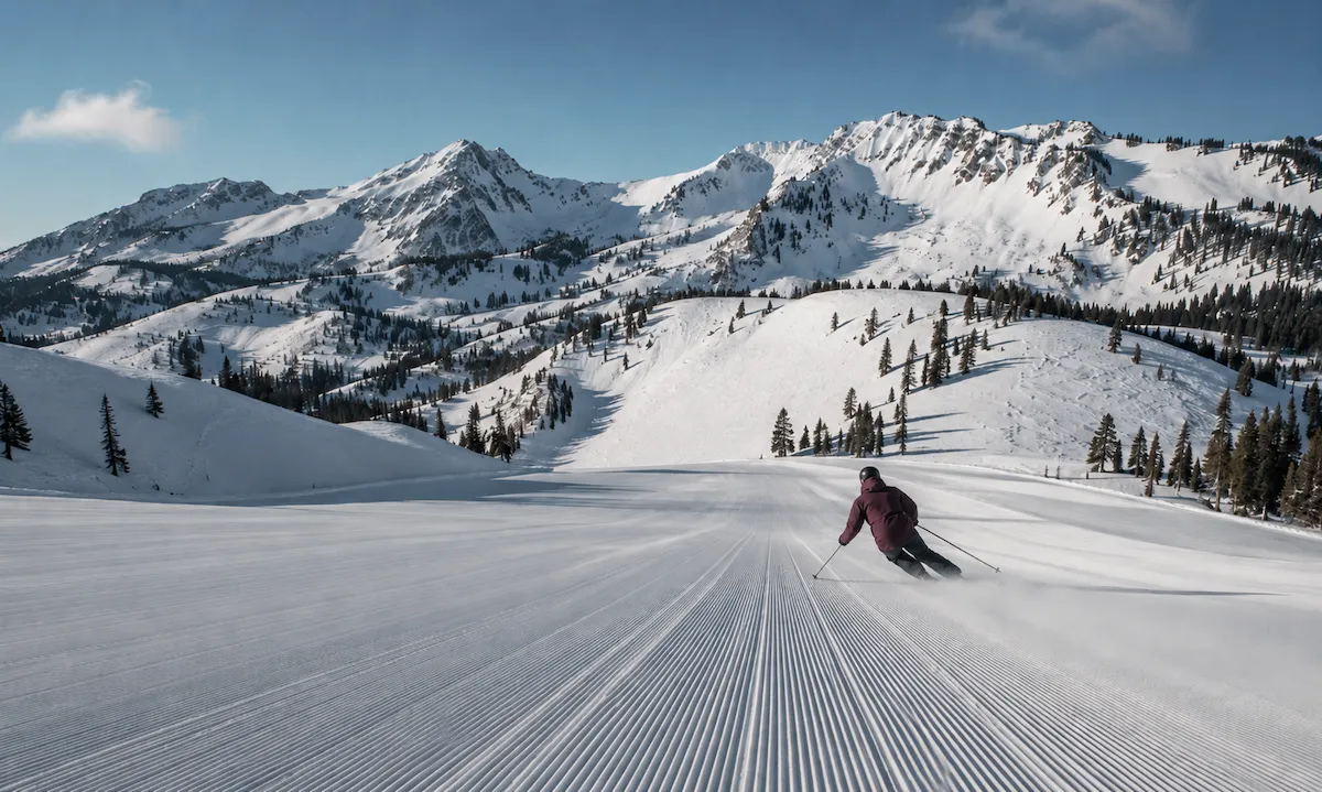 Esquiador descendiendo por una pista amplia y recién pisada en Snowbasin, Utah, con montañas nevadas y terreno olímpico al fondo