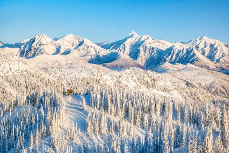 Panorámica soleada de Whitefish Mountain con bosques cubiertos de nieve, montañas escarpadas al fondo y luz dorada de invierno.