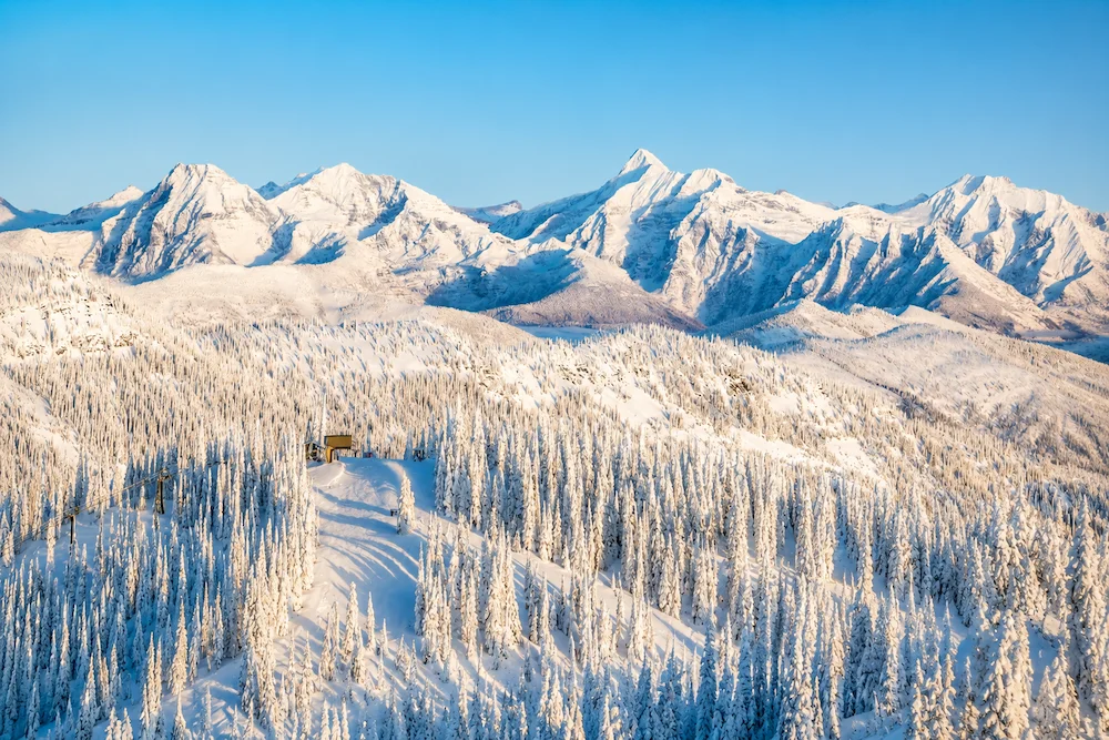 Panorámica soleada de Whitefish Mountain con bosques cubiertos de nieve, montañas escarpadas al fondo y luz dorada de invierno.